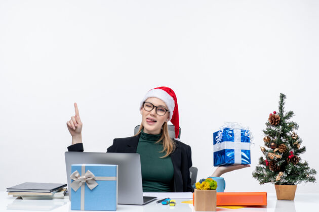 办公室圣诞气氛积极有趣的年轻女子戴着圣诞老人的帽子 戴着眼镜坐在一张桌子上显示礼物指着上面的白色背景笔记本坐着圣诞老人