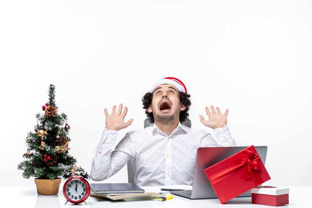 年轻人紧张的年轻商人戴着滑稽的圣诞老人帽在白色背景的办公室里庆祝圣诞节笔记本电脑年轻的商人圣诞节