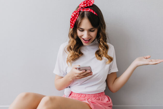 配件一个穿着白色t恤的可爱女孩看着手机屏幕一个飘飘然的微笑着的女士拿着智能手机在家里摆姿势卷发微笑智能手机