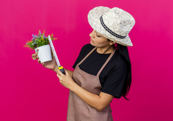 女人年轻漂亮的女园丁戴着围裙和帽子 手里拿着盆栽植物和卷尺 严肃地看着它帽子园丁年轻