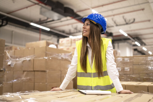 工作勤劳的职业女工人或经理戴着安全帽 穿着反光夹克 靠在大仓库里的纸板箱上向旁边看职业工人安全帽