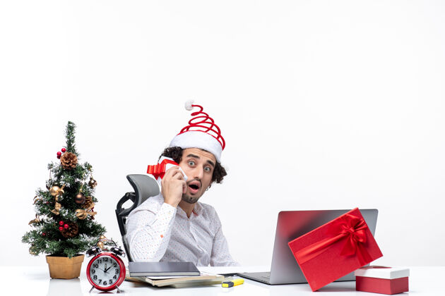 办公室带着圣诞老人帽子 留着胡子的年轻商人举起礼物 看着白色背景上的东西的圣诞节心情电脑看年轻