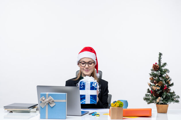 礼物圣诞气氛与戴着圣诞老人帽子和眼镜的年轻女子坐在一张桌子旁举起礼物白色背景年轻女人电脑桌子