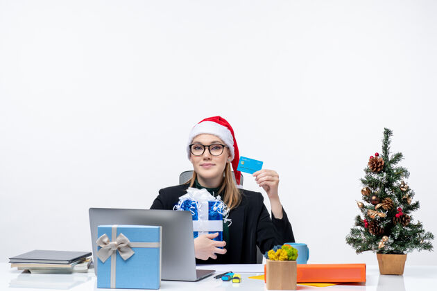 笔记本戴着圣诞老人帽戴着眼镜的快乐年轻女子坐在桌子旁 手里拿着圣诞礼物和白色背景的银行卡办公室抱着银行