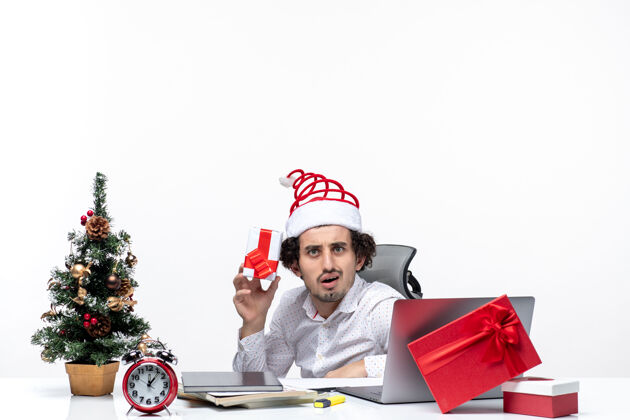 商人带着圣诞老人帽子的年轻商人坐在办公室里 拿着他的礼物 在白色的背景下摆姿势拍照 这是新年的气氛成人礼物相机