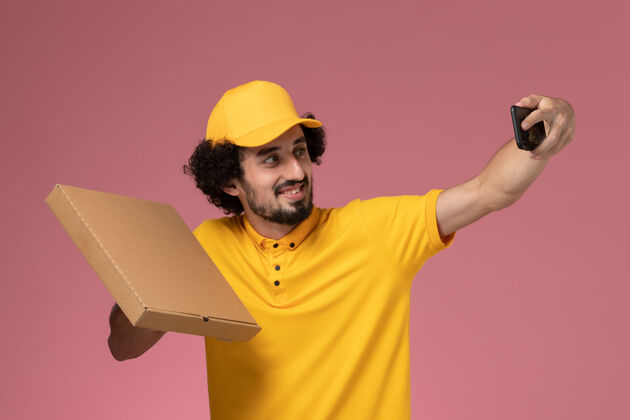 男人正面图身穿黄色制服的男快递员拿着食品快递箱在粉色墙上拍照工作拿着盒子
