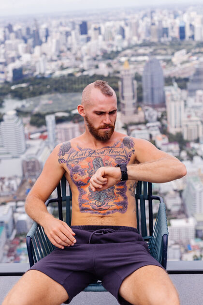 人成功的帅哥 留着胡子 野蛮的纹身 强壮的男人 赤裸上身 戴着手表 坐在高楼的椅子上 城市景色令人惊叹肖像泰国野蛮
