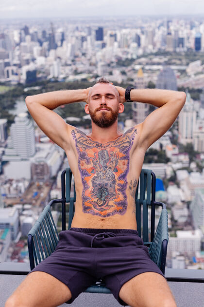 肖像成功的帅哥 留着胡子 野蛮的纹身 强壮的男人 赤裸上身 戴着手表 坐在高楼的椅子上 城市景色令人惊叹野蛮纹身时尚