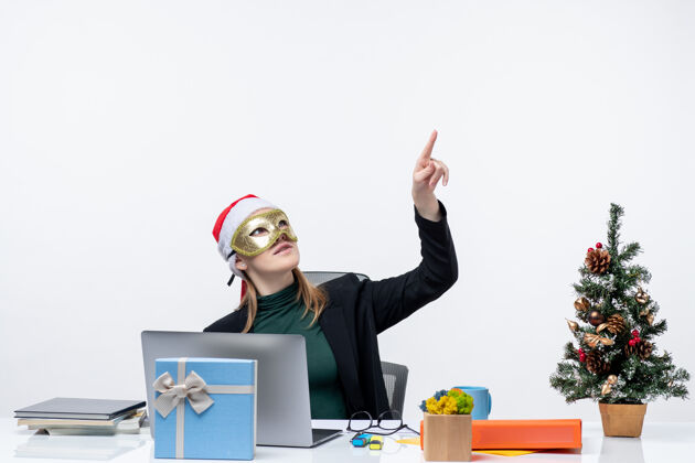 办公室圣诞气氛与紧张的年轻女子戴着圣诞老人的帽子和面具坐在一张桌子上指着白色的背景电脑笔记本指着