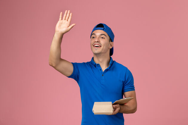 视图正面图身穿蓝色制服披风的年轻男性快递员拿着小快递食品包和笔记本在浅粉色的墙上挥舞年轻男性信使持有快乐