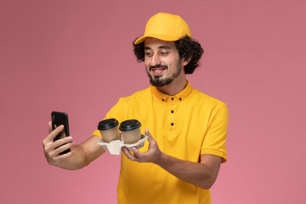 摄影师正面图：身穿黄色制服和披风的男性信使手持棕色咖啡杯 在粉色墙上拍照咖啡拍照人