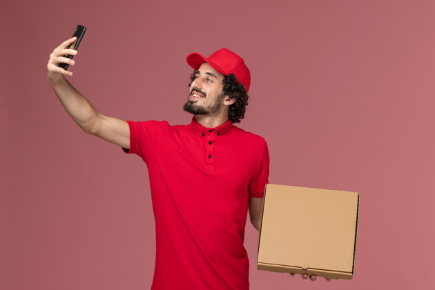 视图正面图穿着红色衬衫和披风的男信使拿着空的食品盒在粉红色的墙上拍照空前面服务