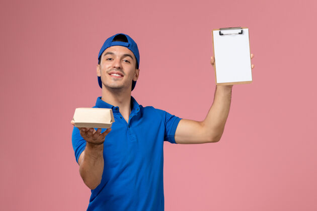 服务正面图身穿蓝色制服披肩的年轻男性快递员手持小快递食品包和笔记本在浅粉色的墙上正面人工人