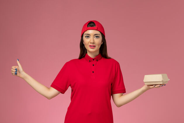 送货正面图：身穿红色制服和斗篷的年轻女快递员 手上拿着一个小小的快递食品包 放在粉红色的墙上食物年轻女快递微笑