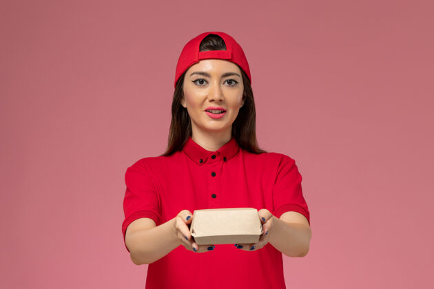 正面正面图：身穿红色制服和斗篷的年轻女快递员 手上拿着一个小小的快递食品包 放在粉红色的墙上送货食物快乐
