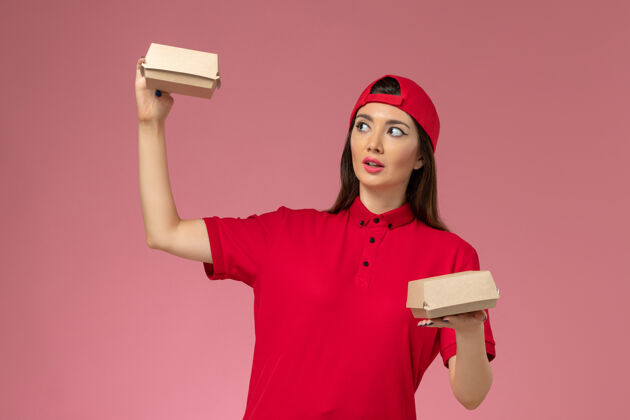 电器正面图：身穿红色制服和披风的年轻女快递员 手上拿着小包裹 手放在粉红色的办公桌上 制服递送送货红色人
