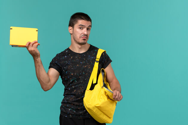 漂亮身穿深色t恤黄色背包的男学生正面图 浅蓝色墙上拿着抄写本男学生教训书