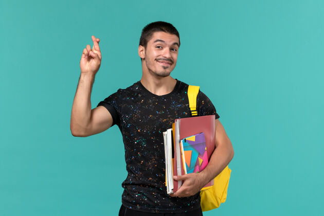 大学身穿深色t恤 背着黄色背包 手里拿着抄写本和文件的男学生正对着蓝色墙壁交叉着手指男学生档案手指