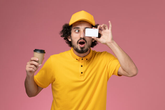 视野正面图：身穿黄色制服 披风的男性快递员手持咖啡杯和粉色墙上的卡片男快递黄色咖啡
