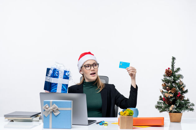 办公室带着圣诞老人帽子戴着眼镜坐在桌子旁的一位积极兴奋的女商人手里拿着圣诞礼物和白色背景的银行卡圣诞老人礼物电脑