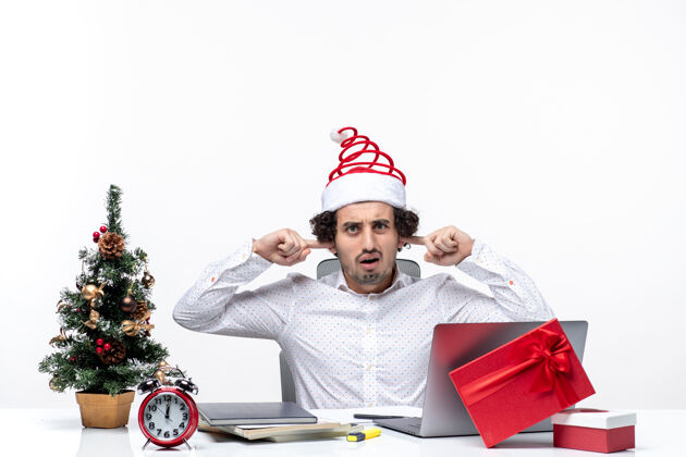 耳朵悲伤的年轻商人戴着滑稽的圣诞老人帽庆祝圣诞节 在白色背景的办公室里 他闭上耳朵不让人听见人年轻人关闭