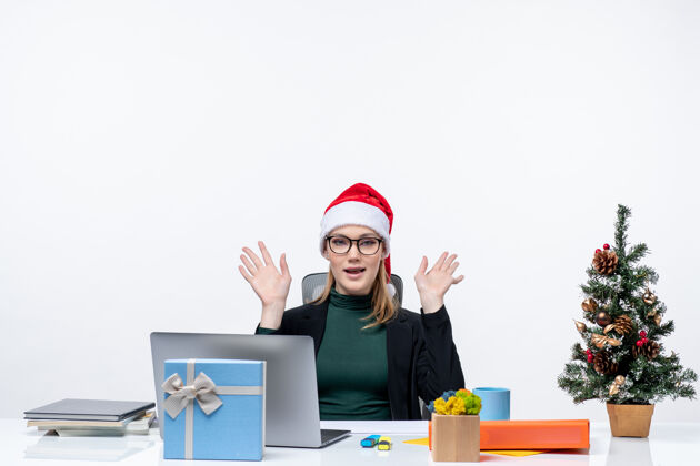 女人新年心情快乐迷人的女人 戴着圣诞老人帽坐在桌子旁 桌上放着一棵圣诞树和一份白色背景的礼物电脑礼物圣诞老人