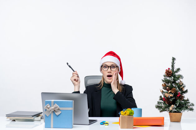 桌子一个不安分的金发女人 戴着圣诞老人的帽子 坐在一张桌子旁 桌子上有一棵圣诞树和一份白色背景的礼物工作圣诞树礼物