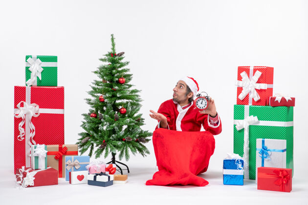 坐着困惑的年轻人坐在地上庆祝新年或圣诞节 拿着礼物和白色背景装饰圣诞树旁的时钟礼物困惑年轻人