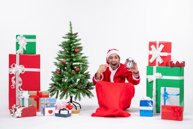 坐快乐的年轻人坐在地上庆祝新年或圣诞节 拿着钟靠近礼物 装饰着白色背景的圣诞树礼物圣诞节年轻人