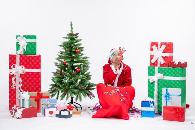 圣诞老人喜庆的节日气氛 有趣的积极的圣诞老人坐在地上玩圣诞装饰品附近的礼物和装饰圣诞树上的白色背景礼物圣诞老人装饰