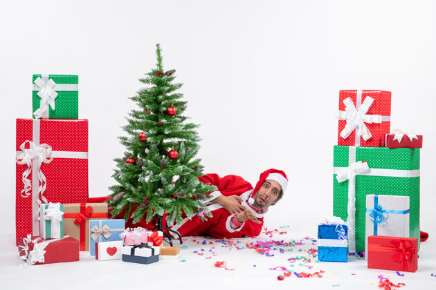 圣诞圣诞老人躺在圣诞树后面 在白色背景上 在不同颜色的礼物旁边 传递着节日的气氛圣诞老人礼物节日