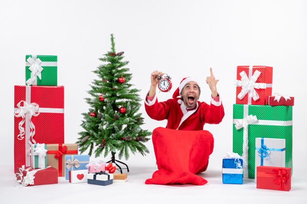 礼物紧张的圣诞老人坐在地上 在礼物和装饰圣诞树旁的白色背景上举起闹钟季节礼物盒子