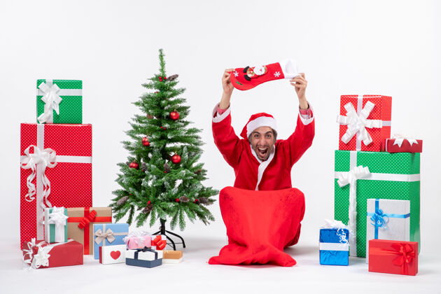 卡通圣诞老人正坐在地上 把圣诞袜举到头上 靠近礼物和装饰在白色背景上的新年树圣诞老人头圣诞树