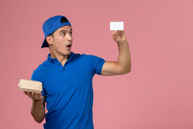 男正面图：身穿蓝色制服披肩的年轻男性信使手拿着小快递食品包和浅粉色墙上的白色卡片生意制服年轻人