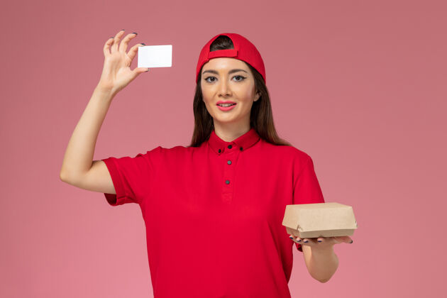 快乐正面图：身穿红色制服斗篷的年轻女快递员 手上拿着小小的快递食品包和卡片 放在浅粉色的墙上生意肖像食物
