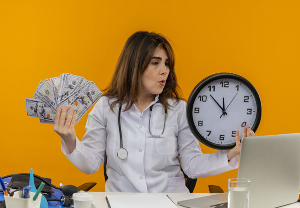 医疗困惑的中年女医生穿着医用长袍 手持听诊器 坐在办公桌旁 拿着笔记本电脑 拿着医疗工具 拿着挂钟 现金放在隔离的橙色墙上 还有复印空间穿着笔记本电脑长袍