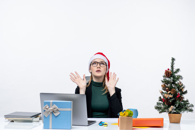 树疲惫的女商人戴着圣诞老人的帽子坐在一张桌子旁 桌子上放着圣诞树和一份礼物 背景是白色的礼物帽子精疲力尽