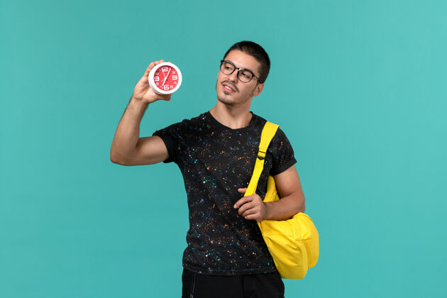 成人身穿深色t恤的男学生正面图黄色背包 蓝色墙上挂着时钟舞会游戏设备背包