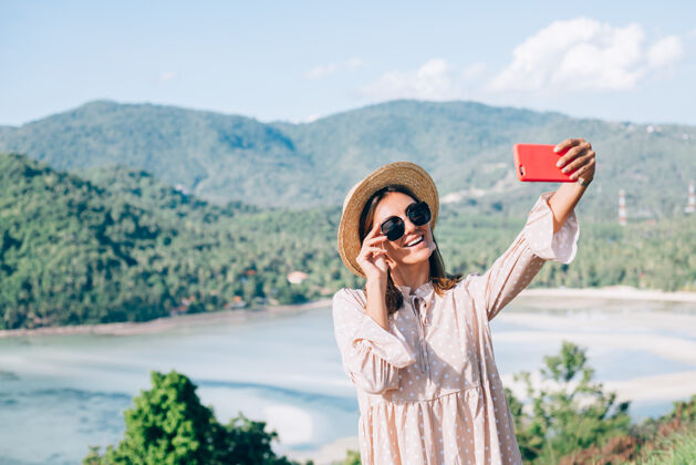 户外一个穿着夏天可爱的裙子 戴着草帽和太阳镜的年轻女人用她的智能手机打电话视频年轻自由