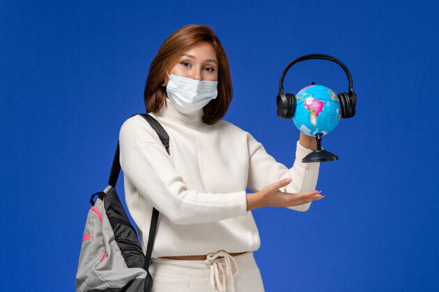 前正面图身穿白色球衣的年轻女学生戴着面具 背着背包 拿着地球仪 蓝色墙上挂着耳机学生地球地球环球