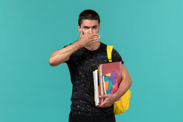 人身穿深色t恤 背着黄色背包 手里拿着抄写本和文件的男学生正对着蓝色的墙上合上嘴教训学院大学