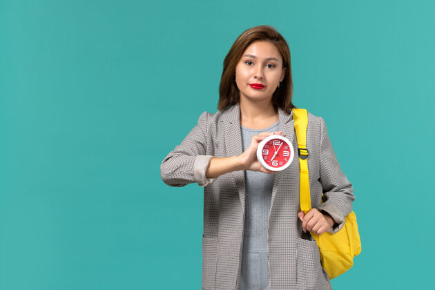 Female身穿灰色夹克 背着黄色背包 在浅蓝色墙上拿着钟的女学生的正视图背包时钟学院