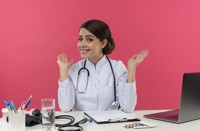 坐着面带微笑的年轻女医生穿着医用长袍 手持听诊器坐在办公桌前 手持医疗工具在电脑上工作 双手张开 留有复印空间听诊器电脑长袍