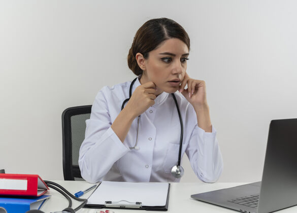 坐着惊讶的年轻女医生穿着医用长袍 手持听诊器坐在办公桌旁 拿着医疗工具坐在电脑上 看着笔记本电脑上的复印空间工具穿着电脑