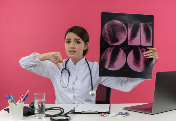 工作悲伤的年轻女医生穿着医用长袍 手持听诊器坐在办公桌旁 拿着医疗工具 拿着x光片 拇指朝下 留着复印空间听诊器电脑医疗
