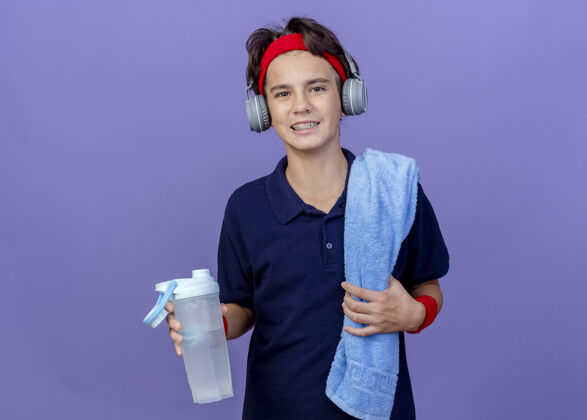 看面带微笑的年轻帅气的运动男孩戴着头带 腕带和耳机 肩上戴着牙套和毛巾 手里拿着水瓶运动牙科抱着
