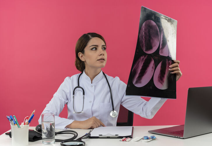工具年轻的女医生穿着医用长袍 手持听诊器 坐在办公桌旁 拿着医疗工具 拿着电脑 看x光片 还有复印空间听诊器工作医疗
