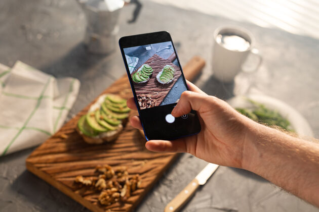 新鲜双手在智能手机上拍下两个美丽健康的酸奶油和鳄梨三明治营养深度块