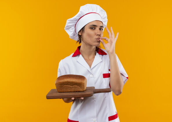 手势年轻的女厨师穿着厨师制服 拿着面包放在砧板上 在隔离的黄色墙壁上展示着美味的手势 还有复制空间看展示板子