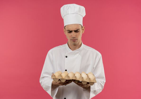 批困惑的年轻男厨师穿着厨师制服 看着孤立的粉红色墙上他手里的一批鸡蛋困惑制服看